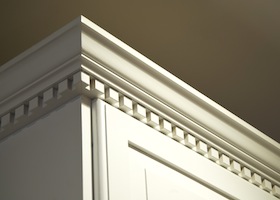 The top crown moulding dentil detail on some creme-color painted solid wood kitchen cabinets. These wall mounted cabinets do not go all the way to the ceiling above them. The lighting is coming from the top right from a ceiling light.